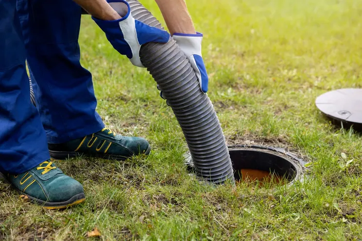 A man wearing blue overalls utilizes a hose to clean a drain, ensuring proper drainage and cleanliness in the area.